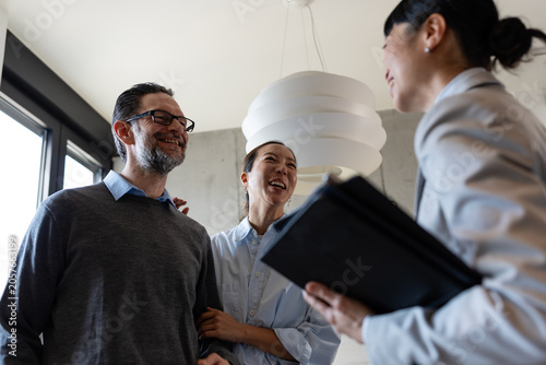 Happy diverse couple laughing with a real estate agent during an apartment tour. Smiling multicultural clients looking at paperwork, discussing a property lease, home purchase, or mortgage deal