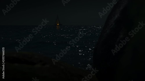Sailboat sailing at night with a faint yellowish light surrounded by darkness with moonlight reflecting in the soft waves