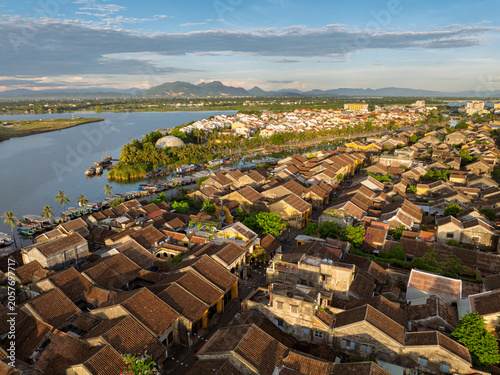Wide aerial panorama of Hoi An rooftops with river scenery and distant mountains.