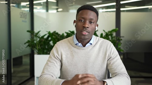 A young african american man in a beige sweater, blue collared shirt and earpiece, uses natural gestures at a white minimalist desk in a bright co-working space, bathed in natural light. Concept of