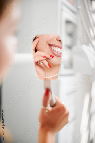 Smiling woman looks at teeth reflection after oral treatment in modern clinic. Dental care concept with mirror and instruments. Good for healthcare campaigns, patient satisfaction, service promotion