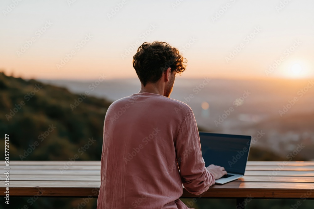 Fototapeta premium Man working on laptop outdoors at sunset on wooden terrace overlooking hills for remote work background and banner