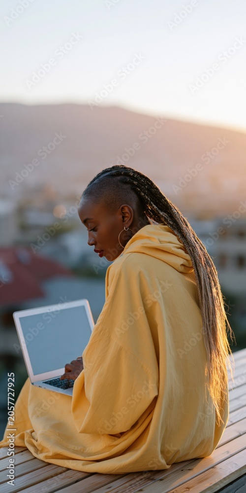 Fototapeta premium Woman in a yellow hoodie seated on a wooden deck using a laptop at sunset with mountain and city views for remote work background and cozy outdoor scene