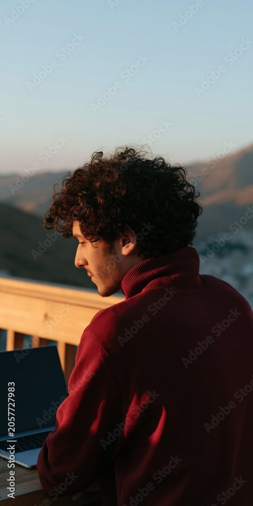 Fototapeta premium Young man working on a laptop on a balcony at golden hour with mountain view for outdoor lifestyle banner and remote work design