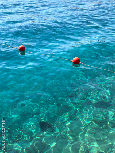 Clear blue water with red buoys floating on a sunny day near a beach or harbor setting. Concept of safe swimming