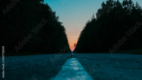 Obraz na plátně Low angle view of an empty asphalt road with a white dividing line running through a dark forest at sunset