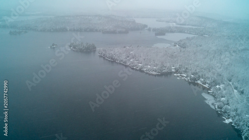Foto A beautiful drone photograph looking down at a vast dark blue lake network with small islands, surrounded by a heavy snow-covered forest shoreline under a hazy overcast winter storm sky