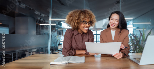 Konstfotografi Two smiling businesswomen collaborate at desk in office, reviewing business tax document at laptop, teamwork, mentoring, workplace communication
