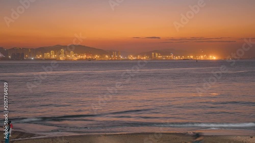 People Watching Sunset Over Calm Sea From Beach With Distant City Skyline