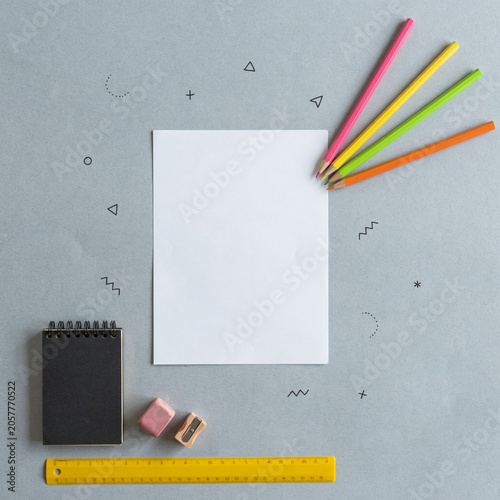 Paper lies flat on a textured surface surrounded by colorful pencils, a ruler, erasers, and a small black notebook.