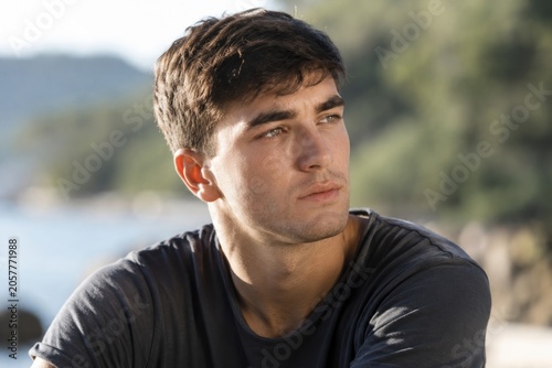 Young male with short brown hair gazes thoughtfully outdoors near a body of water, surrounded by greenery and rocky terrain in a natural setting