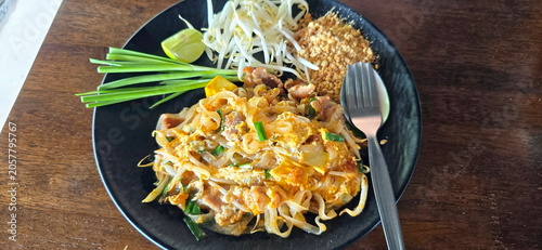 Pad Thai (Pork) served on black plate with cutlery. Side dishes include raw bean sprouts, chives, crushed roasted peanuts, lime wedges. The thin rice noodles are stir-fried in sauce with sour, sweet 
