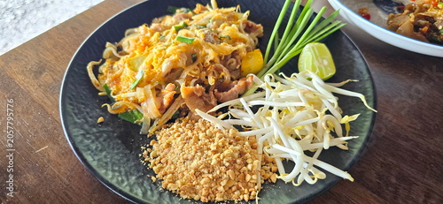 Pad Thai with marinated pork instead of shrimp, served on black plate with spoon and fork. Raw bean sprouts, chives, crushed roasted peanuts, lime wedges. Thin rice noodles stir-fried in a sweet-sour
