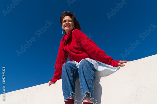 Smiling woman in red sweater sitting on parapet under blue sky