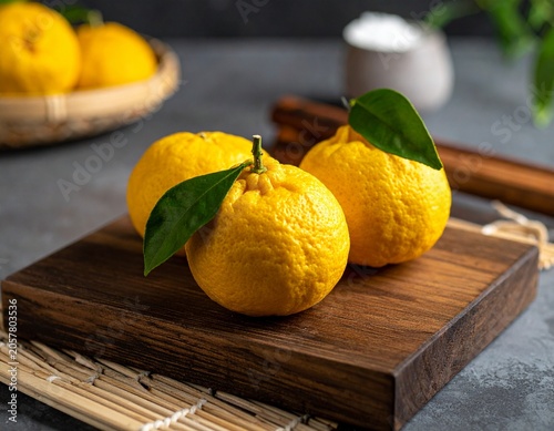 Three bright yellow yuzu fruits with fresh green leaves sit on a dark wooden cutting board, artfully arranged for a still life food photography composition.