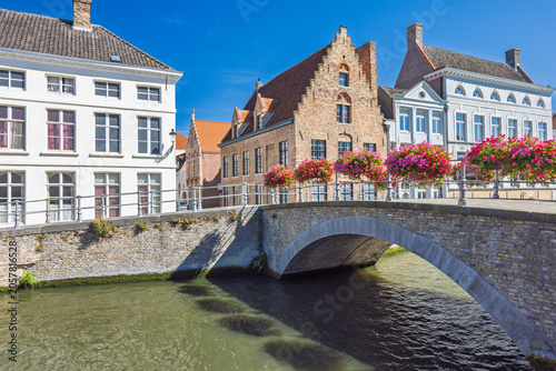 Bruges town in Belgium, Europe. Historic European city canal with a charming stone bridge adorned with vibrant flowers, classic architecture, and clear blue sky on a bright day