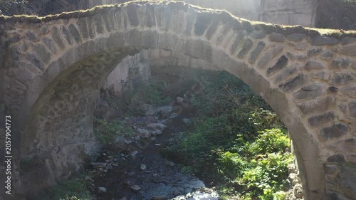 A beautiful historic stone arch bridge stands over a gently flowing river stream surrounded by lush green grass and rocky hillsides on a bright and sunny spring day.

