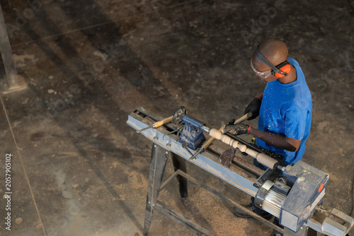 Woodworker is shaping wooden spindle on lathe in workshop on concrete floor with scattered shavings