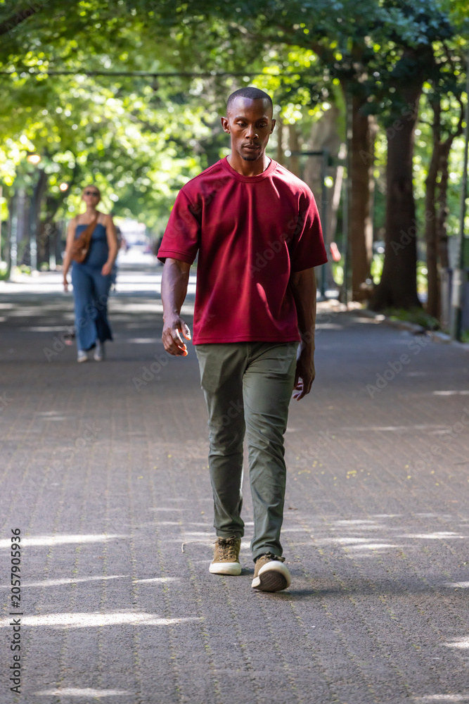 Fototapeta premium African American man walking on paved path under dappled canopy wearing burgundy shirt, olive pants
