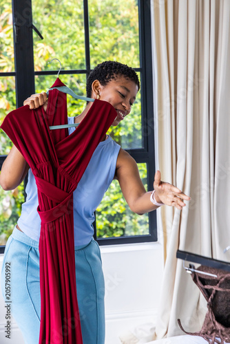 Adult African woman wearing light blue top holding maroon dress by open suitcase at home