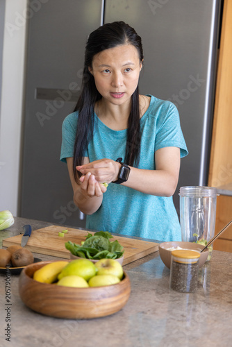 Asian woman in teal shirt smartwatch preparing smoothie in kitchen with cutting board