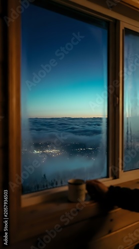 Window view of misty mountain valley at dusk with glowing city lights below, wooden frame interior, serene twilight landscape for travel and lifestyle design projects.