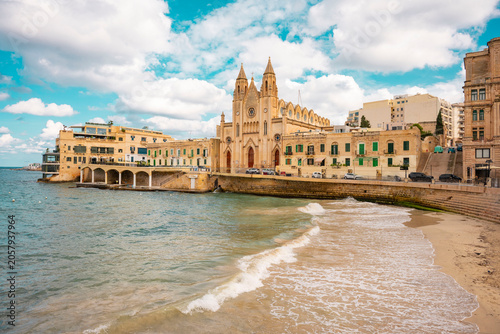This scene depicts Balluta Bay in St. Julian's, Malta, featuring a beach promenade.The large Neo-Gothic building is the Carmelite Church (Parish Church of Our Lady of Mount Carmel).The bay is a popula