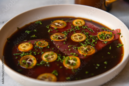 A dish of fresh tuna is served on a white plate. It is garnished with slices of citrus and green herbs. The background shows a restaurant environment, creating a warm dining scene.