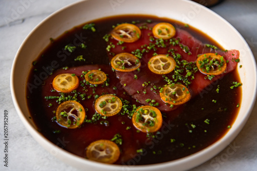 A plate features prepared tuna with thin lemon slices and chopped herbs on a dark sauce. This dish is ready to be served in a restaurant kitchen. The setting shows various textures and colors.