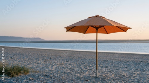 Beach umbrella on sandy shore at sunset, calm water reflecting warm colors, distant hills visible under a clear sky, creating a serene vacation atmosphere