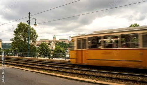 Yellow tram passing through Budapest with Royal Palace visible on hilltop. Motion blur captures iconic Hungarian public transportation with Danube riverbank and historic architecture in background.