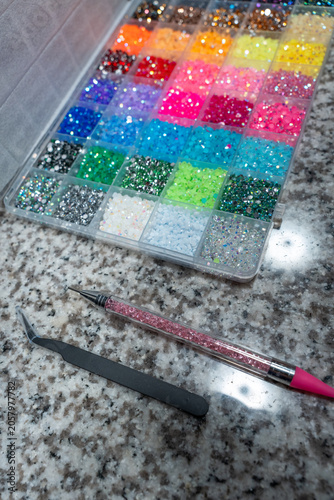 Close-up vertical photo of a box with colorful rhinestones for bedazzling craft. Includes applicator pen and tweezers for precise decoration work. Creative DIY concept, crafting tools, handmade decora