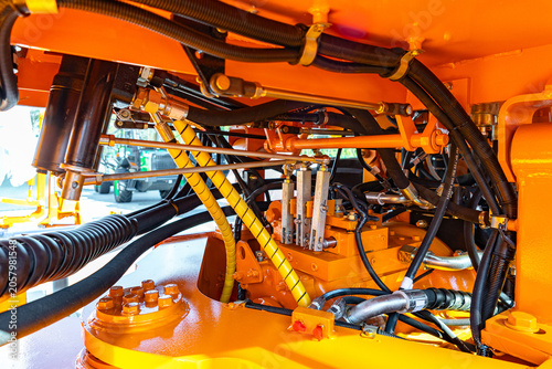 Tight view of yellow painted industrial equipment focusing on hydraulic hoses, metal fittings, bolts and control linkages. Visible are black rubber hoses