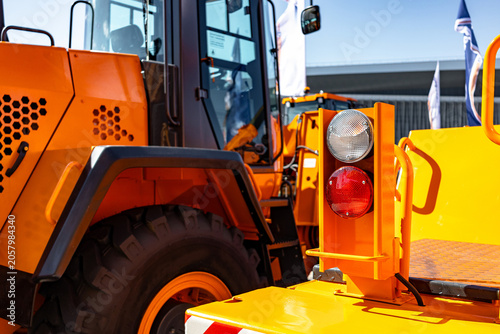 Tight view of the front and side components of a yellow construction vehicle showing metal grille, warning lights, textured paint, and hydraulic fittings