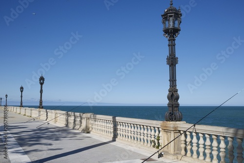 Fishing rods leaning against a decorative stone balustrade overlooking deep blue ocean at Cadiz seafront, Spain. Tall metal street lamps and vivid blue sky.