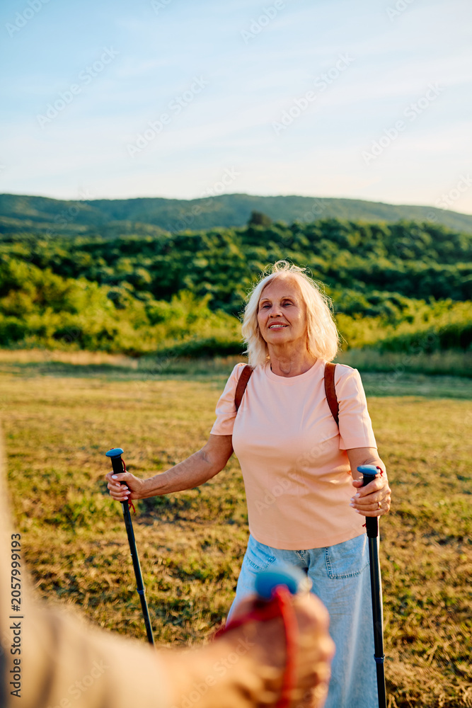 Naklejka premium Happy senior woman with trekking poles enjoying Nordic walking activity in nature, staying active and healthy in retirement