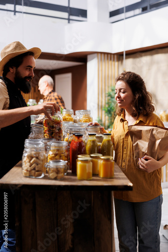 Schilderij op canvas Woman shopping in zero waste grocery store, looking at bulk products in reusable packaging while being assisted by happy storekeeper