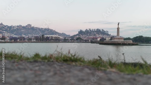Cinematic low-angle ground shot of Lake Anosy and the Monument aux Morts with the Antananarivo skyline in the background during dusk in Madagascar.