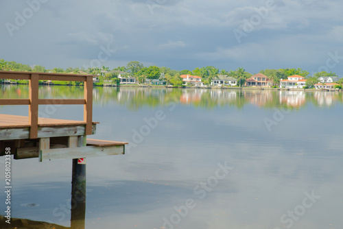 Φωτογραφία Wooden Dock Over Calm Coffeepot Bayou With Cloud Reflections and Bayside Houses Under Stormy Sky in St