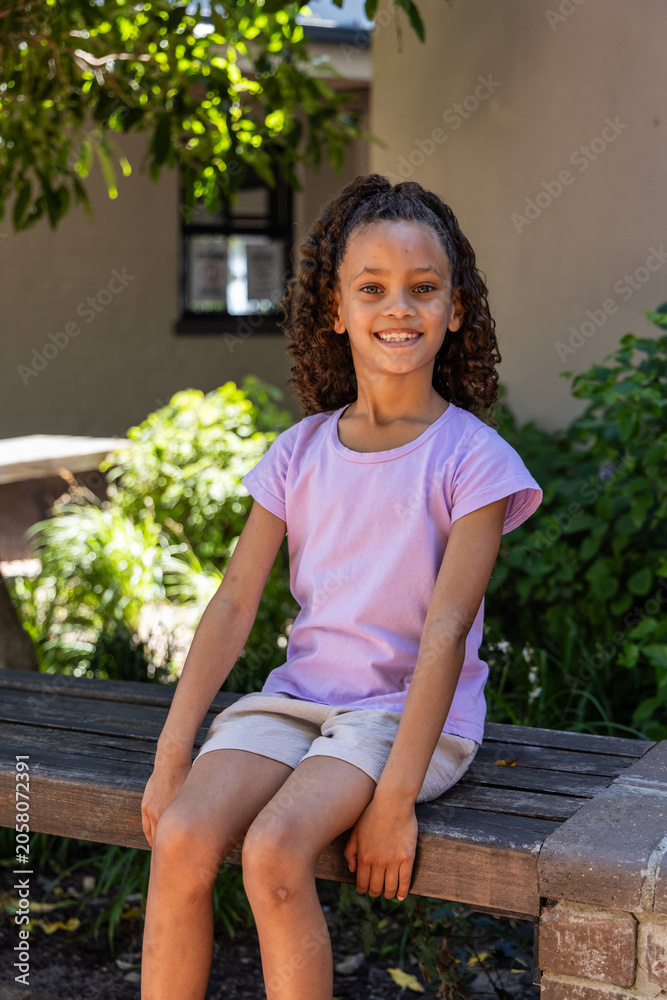 Naklejka premium School-age girl sitting on wooden bench in courtyard, smiling in light purple shirt and shorts
