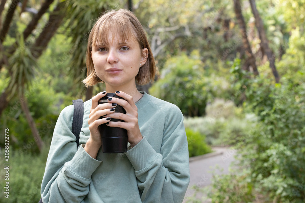 Fototapeta premium Female standing on paved path in park holding black mug, wearing green sweater, backpack strap