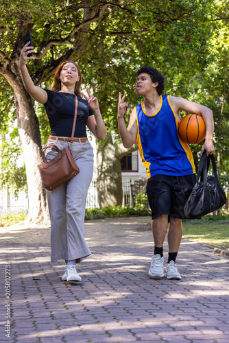African American friends walking park path taking selfie, holding smartphone, basketball, jersey