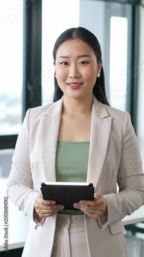 Diverse professional in sharp business attire, subtle smile, direct eye contact, holding a tablet in a high-end, blurred modern office with bright natural light. Concept of professional excellence