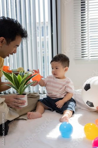 
Warm sunlight streams through blinds. Family of three smiles softly in cozy room. Mother holds infant close, father looks lovingly on. Green plant adds freshness to minimalist shelves
