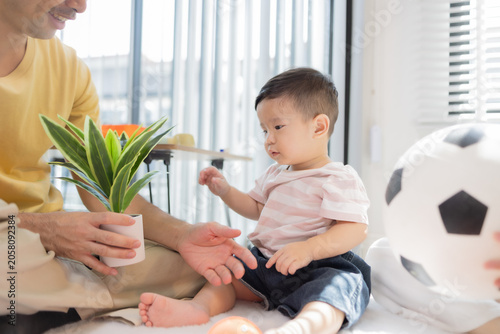 
Warm sunlight streams through blinds. Family of three smiles softly in cozy room. Mother holds infant close, father looks lovingly on. Green plant adds freshness to minimalist shelves
