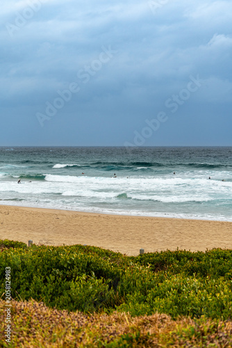Surfers waiting for waves at a sandy beach under cloudy skies