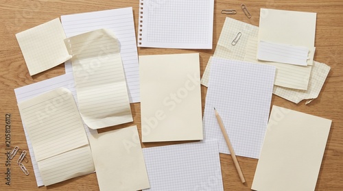 Top view of scattered blank papers, notes, notebook, and a pencil on wooden table.