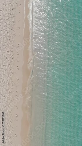 Extreme high-angle aerial video of pristine tropical beach. Turquoise ocean water gently laps light golden sand, reflecting bright sunlight. Minimalist, expansive negative space for text, idyllic