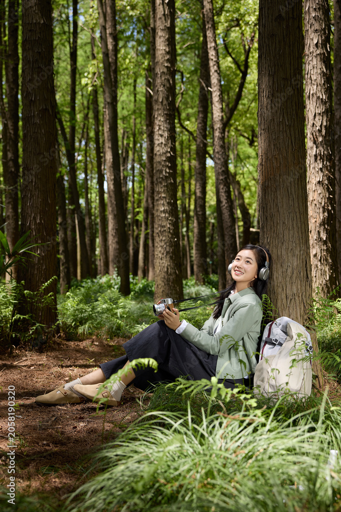 Fototapeta premium Young woman with camera and headphones resting under tree in forest