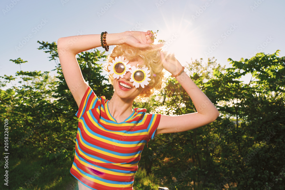 Obraz premium Cheerful woman outdoors, wearing a striped shirt and sunflower sunglasses, basking in warm sunshine, carefree pose, vibrant colors, lush greenery background, joyful spontaneous moment, lively